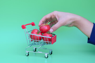 Female hands hold red ripe tomato cherry near little shopping trolley full of tomatoes on green background.Concept of choosing and buying vegetables in supermarket.