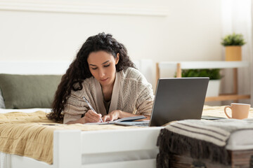 Focused young woman using laptop, working or studying online, taking notes during business meeting or webinar at home