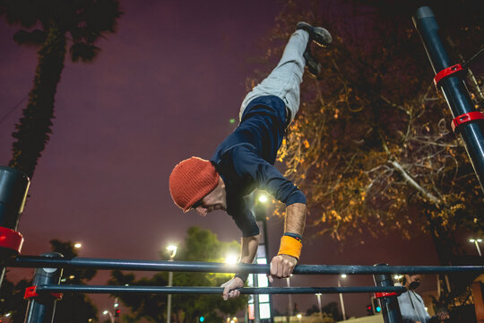 Healthy and fit young latin man with wool cap and wristbands doing calisthenics (inverted pose on high bars) on a street workout park with beautiful and colorful sunset sky