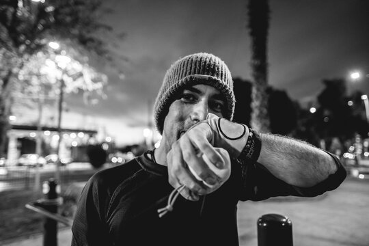 Portrait of a smiling, healthy, handsome, and fit young latin man with wool cap and wristbands holding a bag of magnesium on a street workout park with beautiful sunset sky (in black and white)