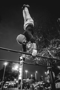 Healthy and fit young latin man with wool cap and wristbands doing calisthenics (inverted pose on high bars) on a street workout park with beautiful and colorful sunset sky (in black and white)