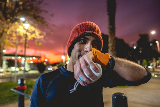 Portrait of a smiling, healthy, handsome, and fit young latin man with wool cap and wristbands holding a bag of magnesium on a street workout park with beautiful and colorful sunset sky