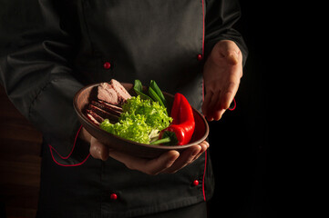 The chef is holding a plate with sliced steak and vegetables. Cooking concept on dark background with advertising space