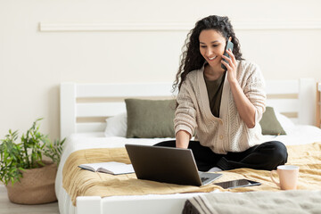 Smiling young woman talking on cellphone, using laptop, sitting on bed at home, empty space