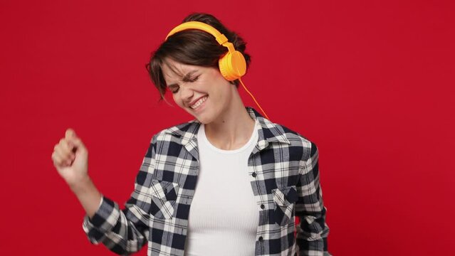 Vivid cheerful woman 20s she wearing basic white t-shirt checkered shirt listen music in headphones dance fooling around have fun enjoy relax isolated on plain solid dark red color background studio