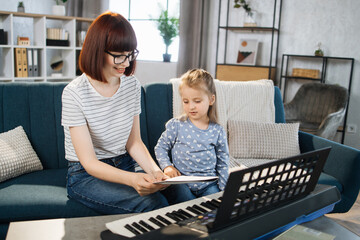 Child girl learning to play piano using notes together with her mother. Remote learning from home....