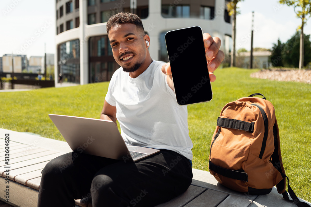 Poster happy black guy with laptop showing smartphone with blank screen, recommending mobile app or website