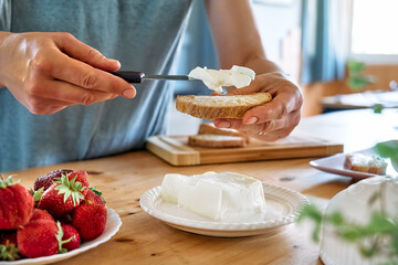 Woman making summer strawberry sandwich. Female hands spread stracchino cheese on bread for toast....
