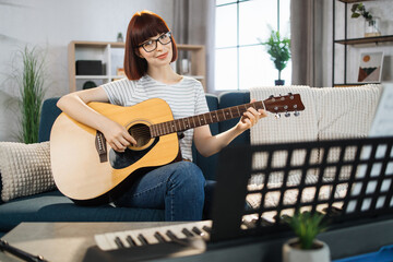 Young woman plays the guitar on bright living room at home. Girl sitting on sofa with a musical...