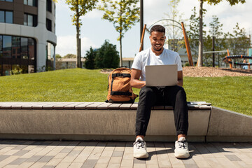 Distance education concept. Black male student with laptop sitting outdoors and watching educational webinar