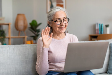 Glad european senior lady with gray hair in glasses waves her hand, looks at computer in living room interior