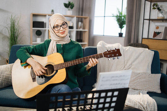 Portrait of young arab female with hijab playing on guitar. Pretty muslim girl learning to play guitar at home. Beautiful student woman playing musical instrument.