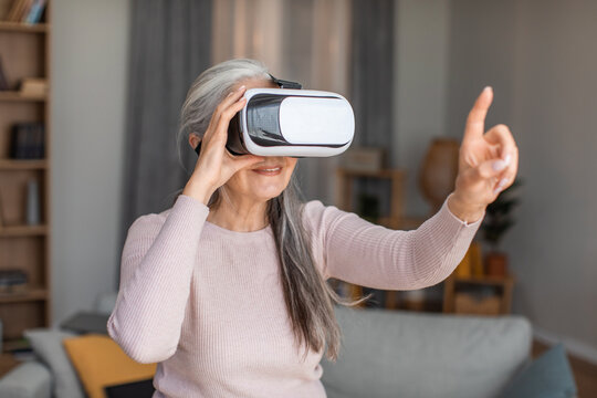 Cheerful european senior woman with gray hair in vr glasses plays in online game at free time in room interior