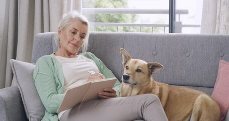 Senior woman writing notes in journal, playing crossword puzzle, sitting on sofa with pet dog. Elderly lady relaxing in home living room, bonding with mix breed Africanis canine. Mans best friend - Powered by Adobe