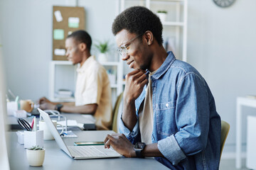 Side view portrait of smiling black man using laptop computer while working in IT office, copy space