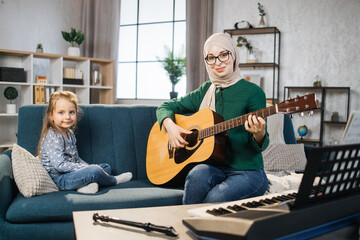 Little cute girl with music muslim teacher having lesson at guitar at school of music. Smiling young arab woman looking at small kid girl sitting on the sofa and playing guitar together in classroom.