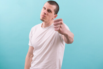 An angry man in a white t-shirt shows his fist on a blue background. Fight. Gesture. Threat. Boxing. Camera. Caucasian. Hand. Arm. Attack. Emotional. Evil. Expression. Force. Fury