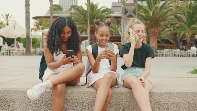 Three pre-teenage girls friends are sitting on the waterfront using mobile phone. Three teenagers watching videos, photos, making calls on the outdoors in urban cityscape background