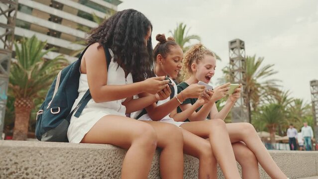 Three girls friends pre-teenage are sitting on the waterfront using mobile phone. Three teenagers playing games on a smartphone on the outdoors in urban cityscape background