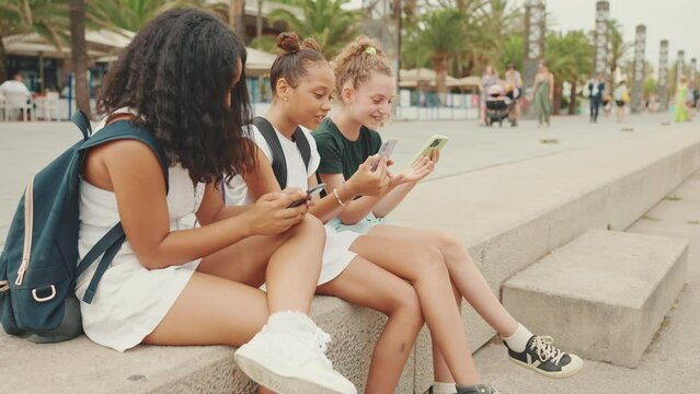 Three pre-teenage girls friends are sitting on the waterfront using mobile phone. Teenagers watching videos, photos, posting on social networks on the outdoors in urban citiscape background