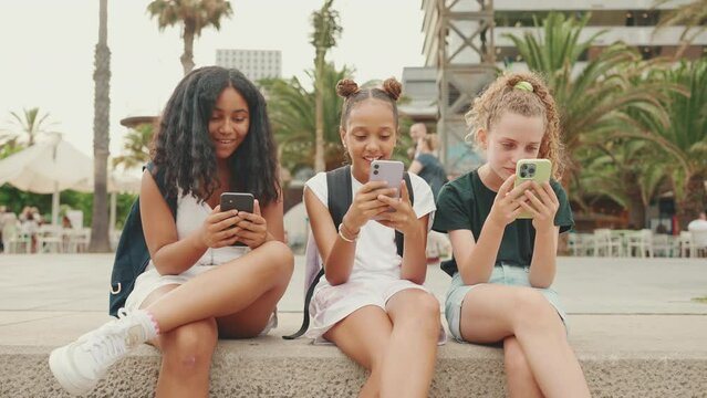 Three pre-teenage girls friends are sitting on the waterfront using mobile phone. Teenagers are watching videos, photos, posting on social networks on the outdoors in urban citiscape background