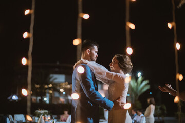 Outdoor wedding party. The first dance of the newlyweds. The bride and groom are dancing in the lights of garlands at night