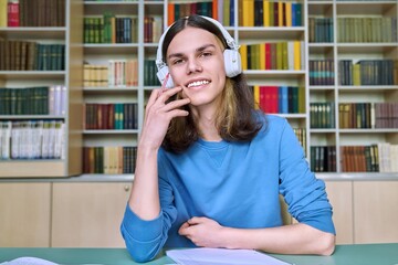 Teenage college student in headphones looking at webcam, sitting at desk in library