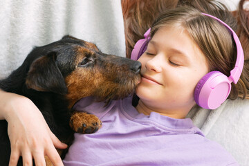 Happy teenage girl in lilac t-shirt in pink headphones listens to music and has fun playing with dog