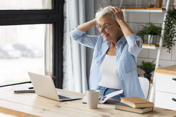 Attractive senior woman working at home in the kitchen using her laptop drinking tea. Concept of mature female using internet and technology for learning, shopping, bank services, freelance job