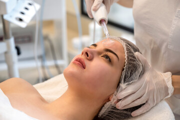 Obraz na płótnie Canvas Young woman receiving electric darsonval facial massage treatment at beauty salon.