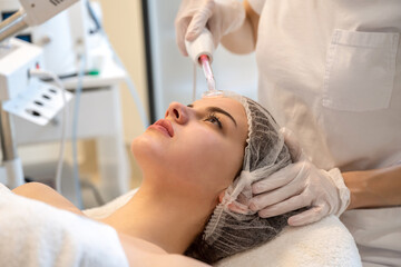 Beautician applying electric Darsonval facial massage treatment with special tool over patients face in beauty salon
