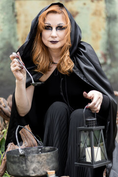 Attractive beautiful woman in a Halloween costume black cloak and scary make-up. Young girl in a look of a witch dressed up for a party making fairy potion holding black lantern