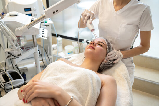 Beautician applying electric Darsonval facial massage treatment with special tool over female patients forehead in beauty salon