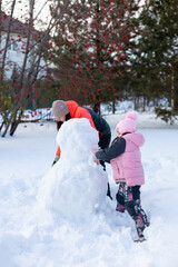 Woman with little daughter on snow making snowman with hands together in evening with rowan and fir trees and fence in background. Winter family time outside.