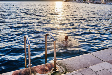 The boy jumps into the sea from a stone pier at sunset