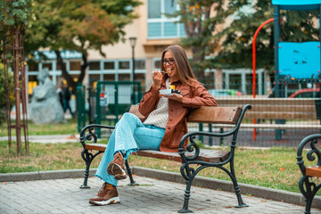 A girl wearing glasses and fashionable clothes sits on a park bench and eats delicious sweets. Selective focus.