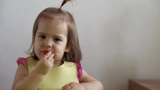 Close up of lovely baby girl eating have breakfast fresh raw vegetables sour tomato by herself at home. Adorable possitive happy toddler child eats dirty face at table. Food, nutrition healthy life