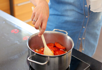 a woman cooking italian cuisine, making tomato soup or sauce for pasta, detailed view of hands holding wooden spoon stir fried chopped tomatoes in a pot on electric stove, background for text.