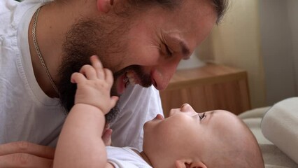 Authentic Bearded Long-haired Young Neo Father And Newborn Baby Looking Each Other Smiling On Bed. Dad Laying With Infant Child. Children, Parenthood, Childhood, Life, Love, Fatherhood, Family Concept