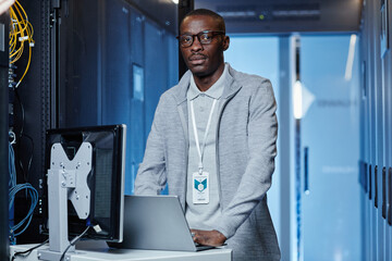 Waist up portrait of adult black man working as IT engineer in server room and looking at camera