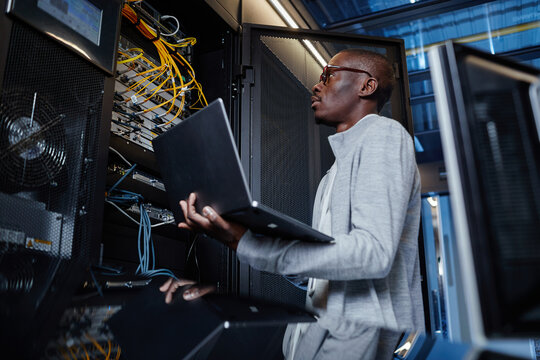 Side view portrait of black man working as IT engineer holding laptop while setting up internet network in server room - Powered by Adobe