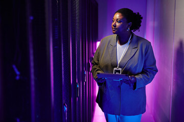 Portrait of female system administrator inspecting data network in server room lit by neon light,...