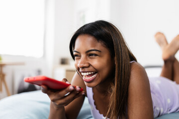 Happy teenager african american girl sending audio message to her friends on mobile phone app while relaxing on bed at home