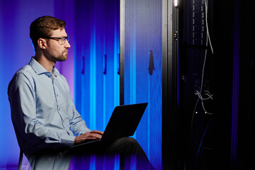 Side view portrait of network engineer using laptop in dark server room lit by neon light, copy...