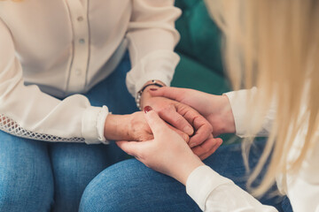 Close-up inner shot of two unrecognizable women sitting on a green couch holding their hands, supporting each other. High quality photo