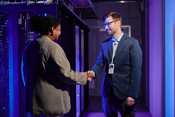 Waist up portrait of two business partners shaking hands while standing in data center interior