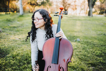 Young brunette woman with glasses holding a cello at sunset in the park, on a green grass. Copy...