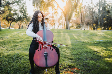 Young brunette woman with glasses playing cello at sunset in the park, on a green grass. Copy space. © padnob