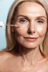 Fototapeta Close up of middle aged woman face, using dropper tool to apply C-Serum moisturizer, collagen on her skin, smiling, standing over blue background obraz