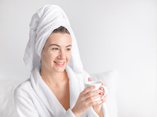 a beautiful young woman in a bathrobe and with  bath towel on her head sits on the bed and holds a...